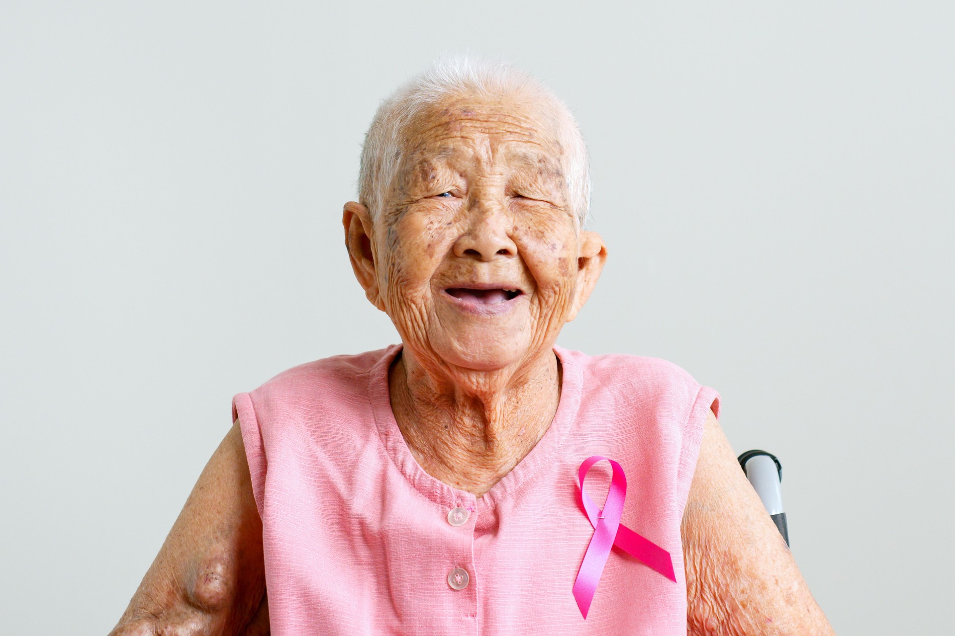 Portrait of senior Asian woman in pink shirt with pink ribbon smiling on gray background for October breast cancer awareness month
