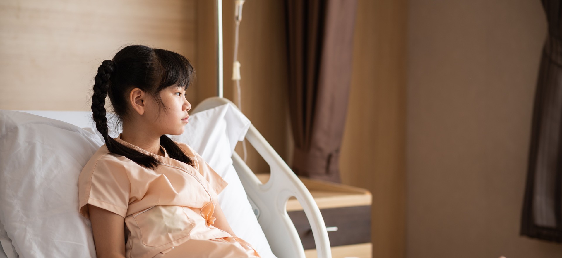 Sick Asian little girl sitting on hospital bed feeling sad and unhappy. Homesick girl looking hopefully outside and want to go home soon while being treated in the hospital. Banner ratio photo.